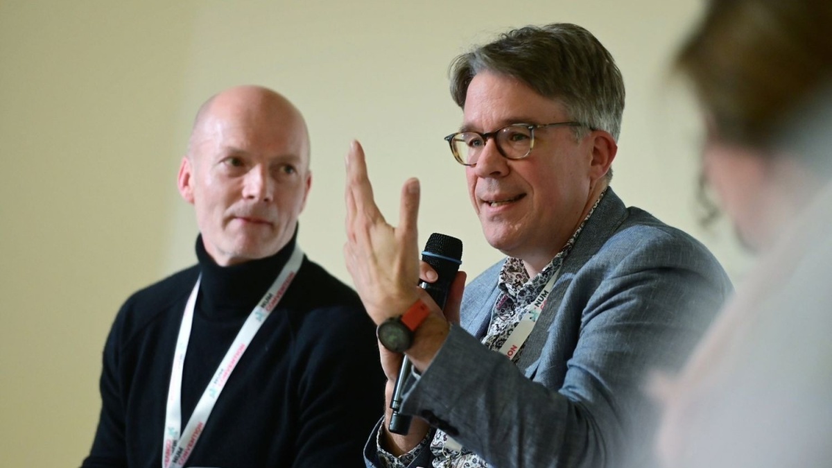 Zwei Personen sitzen nebeneinander. Die Person rechts spricht in ein Handmikrofon und gestikuliert mit der freien Hand, während die Person links aufmerksam zuhört. Beide tragen Businesskleidung und Namensbänder. Im Vordergrund sind unscharf weitere Personen zu erkennen.