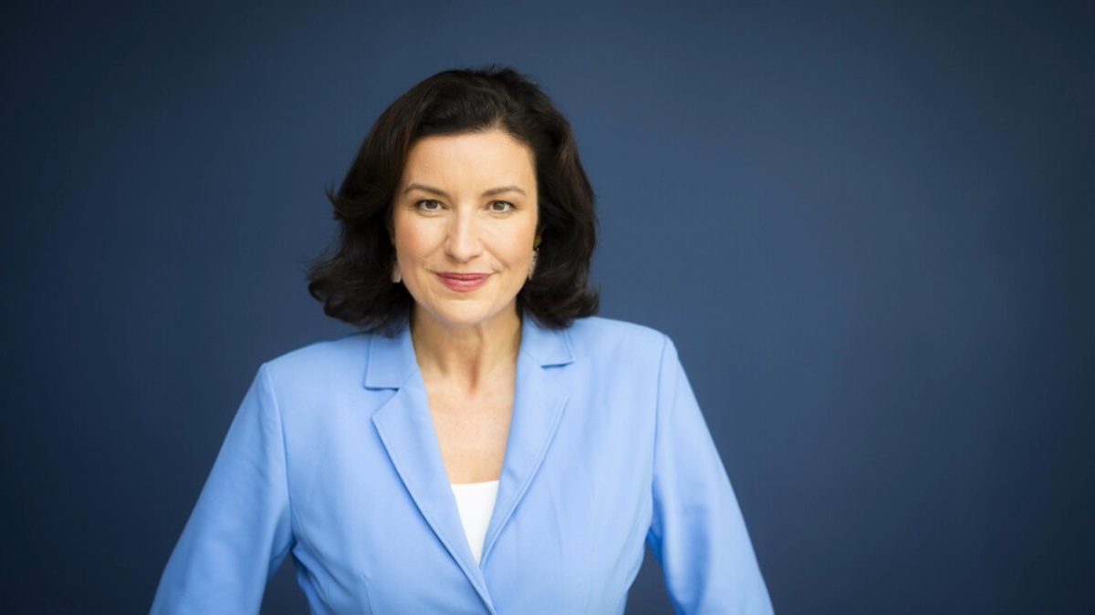 Porträt einer Frau mit schulterlangem dunklem Haar, die direkt in die Kamera blickt. Sie trägt ein hellblaues Sakko über einem weißen Oberteil und steht vor einem einfarbig dunkelblauen Hintergrund.