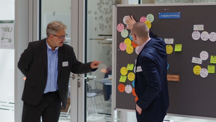 Zwei Männer stehen vor einer Pinnwand, auf der mehrere bunte, beschriebene Zettel angepinnt sind