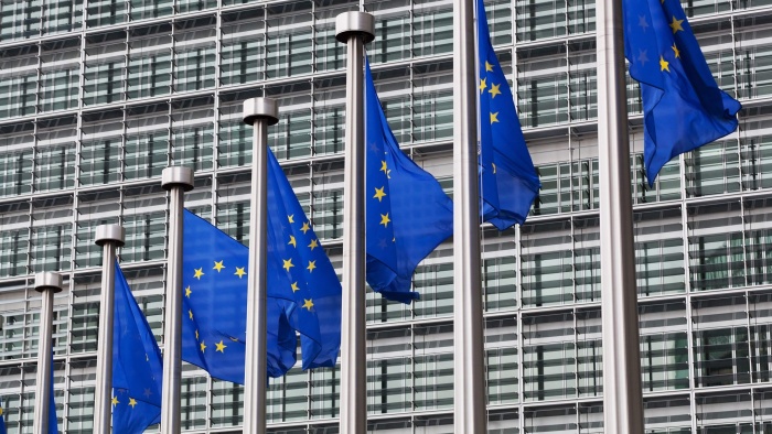 Several European Union flags are flying in front of the glass façade of a modern EU building.