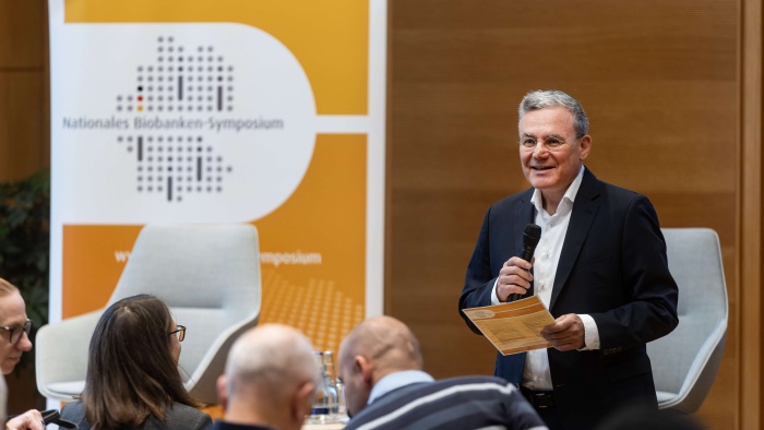 Ein Mann im dunklen Anzug steht mit Mikrofon und Unterlagen vor einem Publikum und spricht auf dem Nationalen Biobanken-Symposium. Im Hintergrund sind zwei helle Sessel sowie ein orange-weißes Veranstaltungsbanner mit dem Logo des Symposiums zu sehen.
